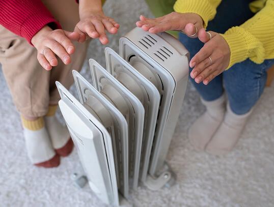 Two pairs of hands reach toward a portable heater, providing warmth in a cozy indoor setting. The scene emphasizes comfort and warmth during colder weather.