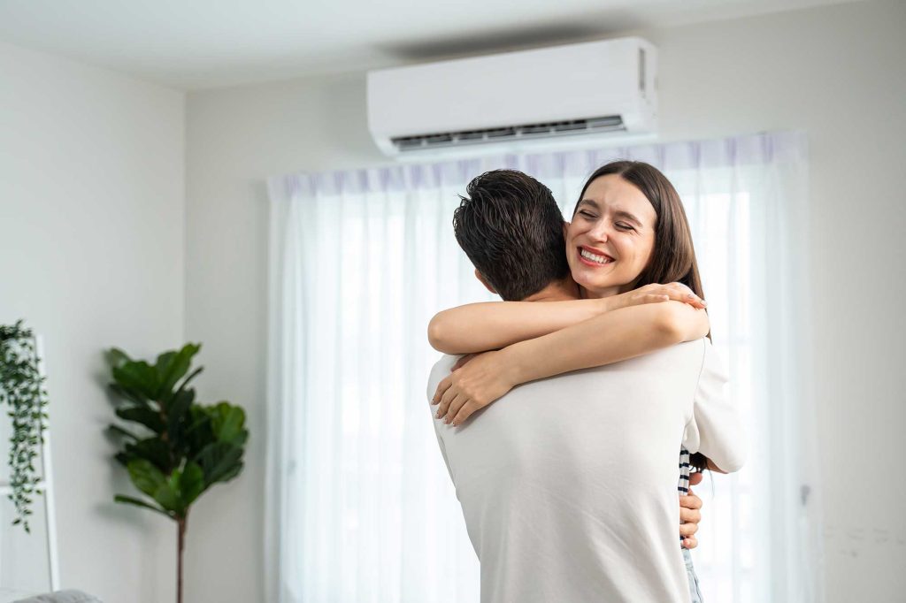 A woman joyfully hugs a man in a bright room with white curtains and an air conditioner above. A leafy plant adds a touch of greenery, creating a cozy atmosphere.