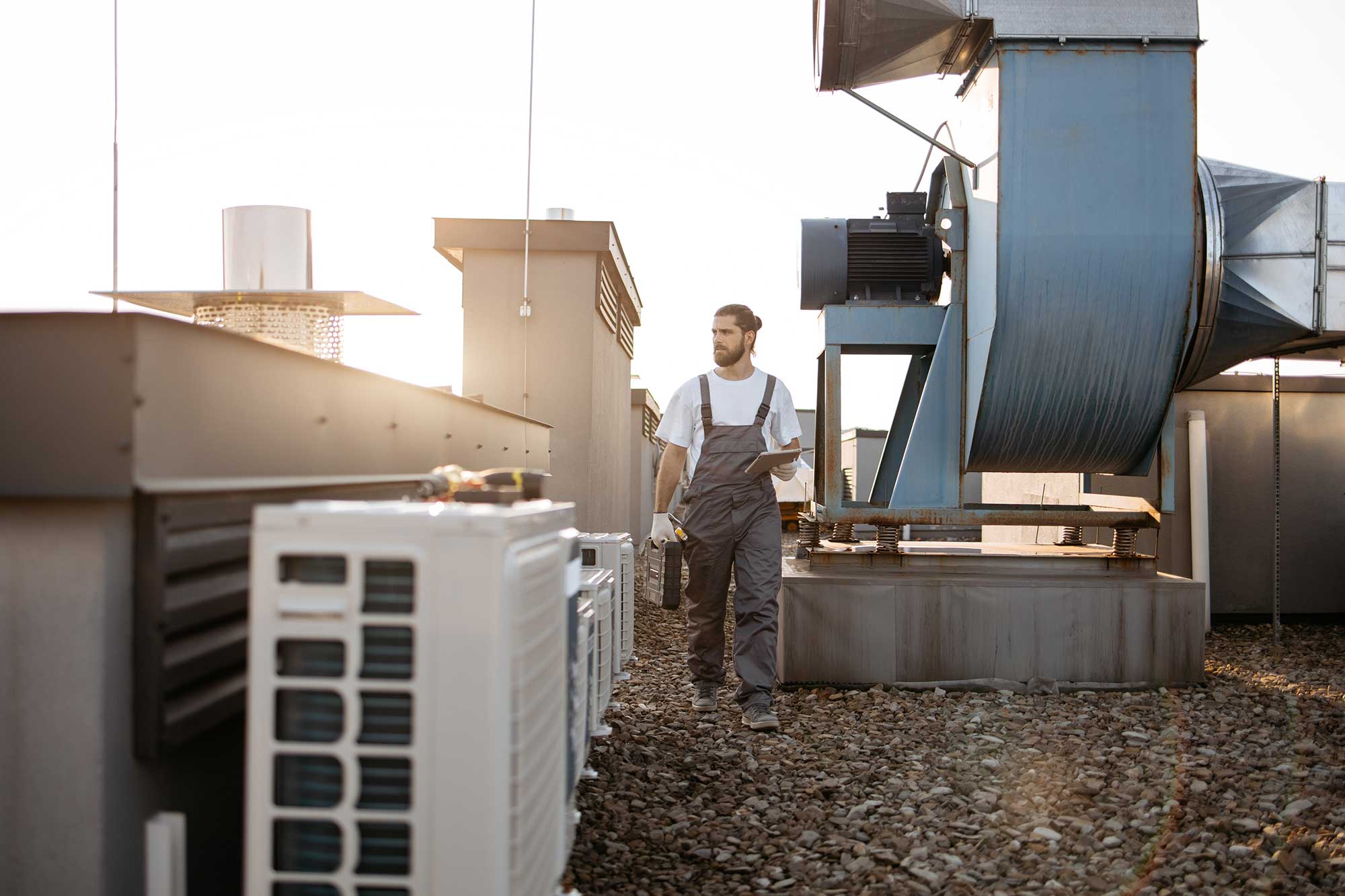 Technician in overalls walking on a rooftop with HVAC units, carrying a toolbox and a clipboard.