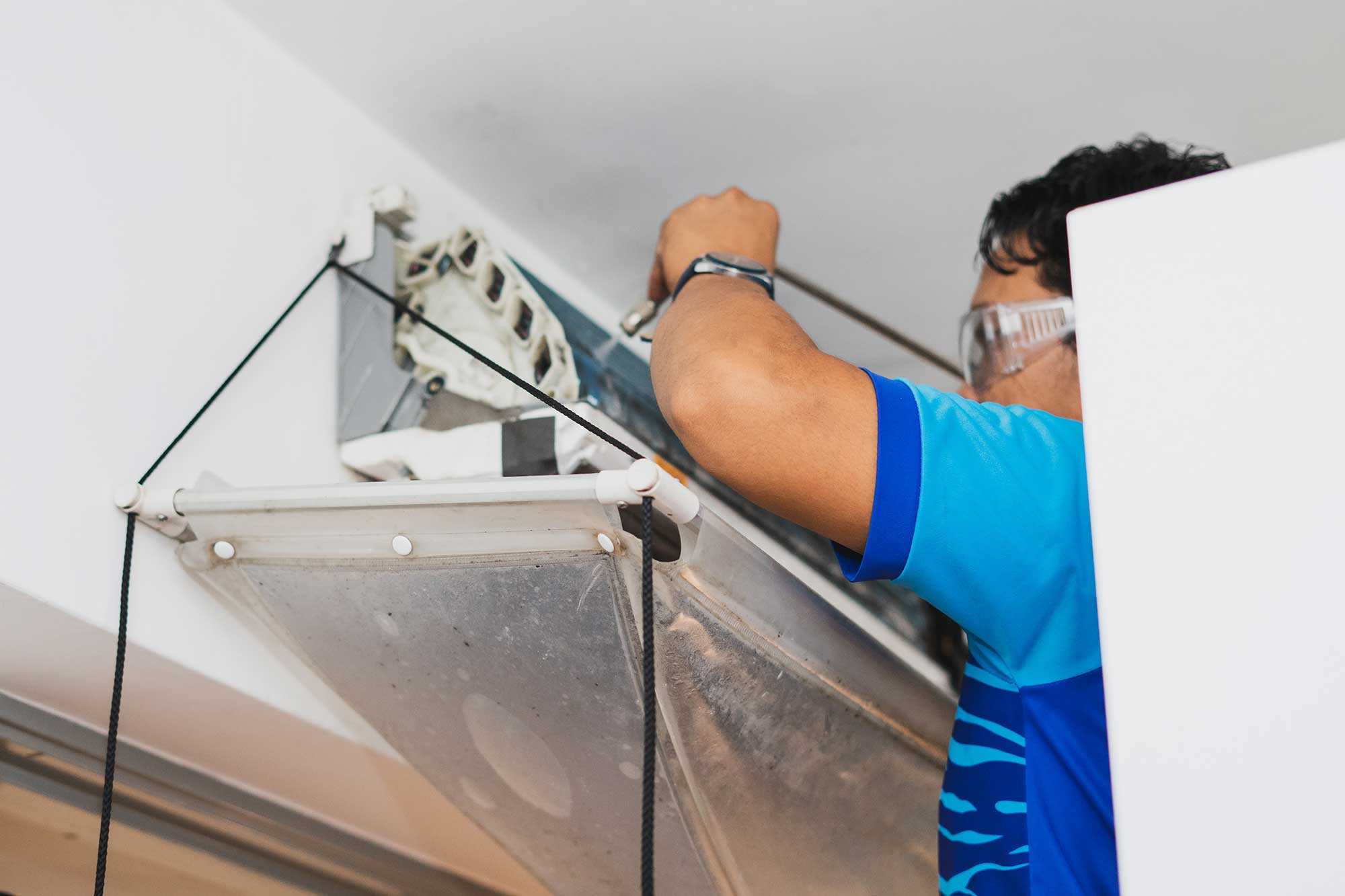 A technician in safety goggles and a blue shirt services a wall-mounted air conditioner, adjusting wires with a focused expression.
