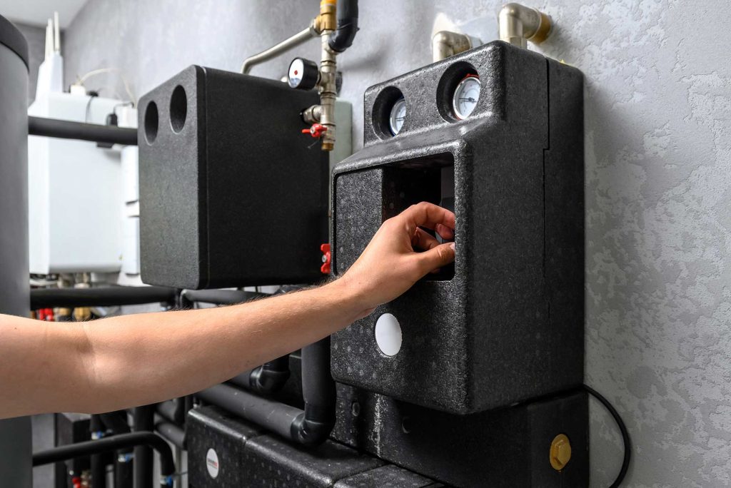 Hand adjusting a gauge or valve on a black insulated heating system with pipes and pressure meters against a textured gray wall.