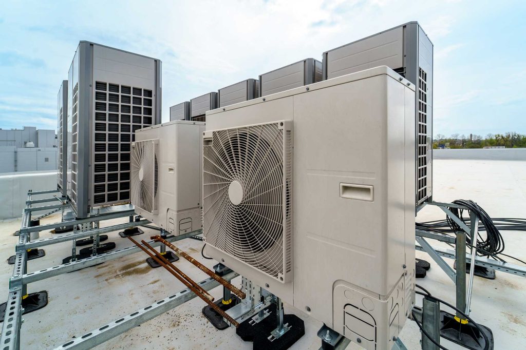 Air conditioning units on a rooftop, aligned in a row under a blue sky. Metal frames support the units, with cables and pipes visible, suggesting an industrial setting.