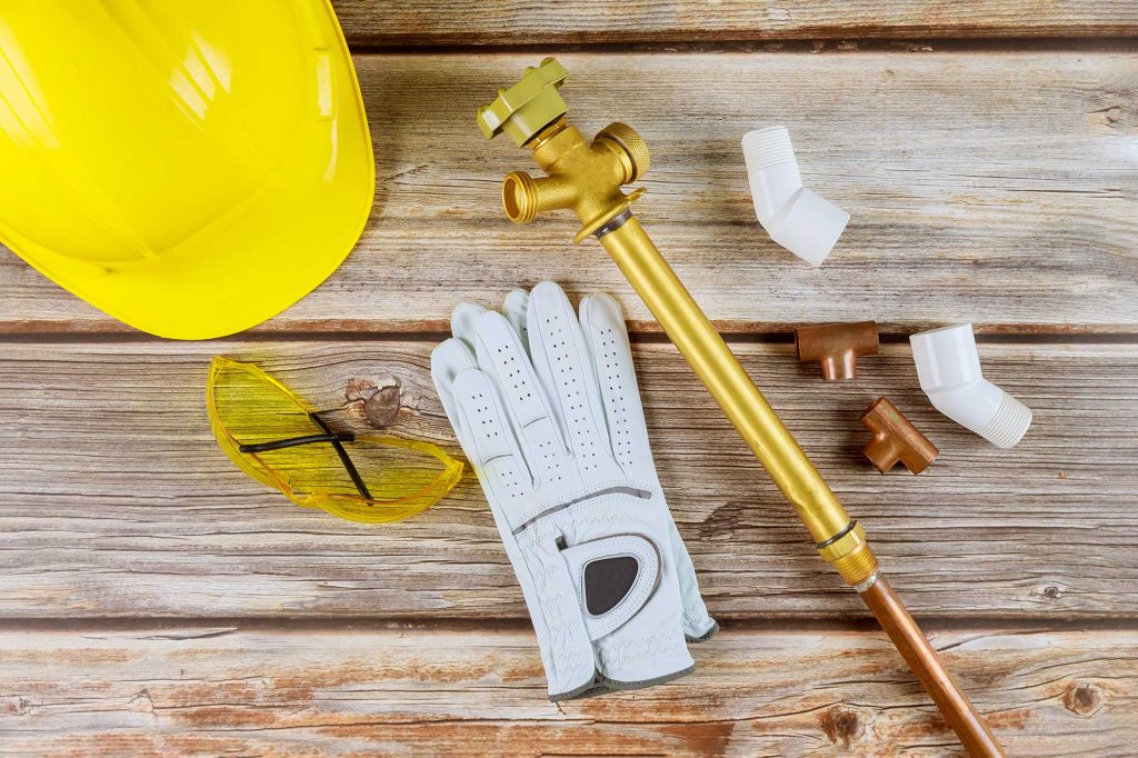 Yellow hard hat, safety glasses, white gloves, brass valve, and assorted pipe fittings arranged on a wooden surface, suggesting plumbing or construction work.