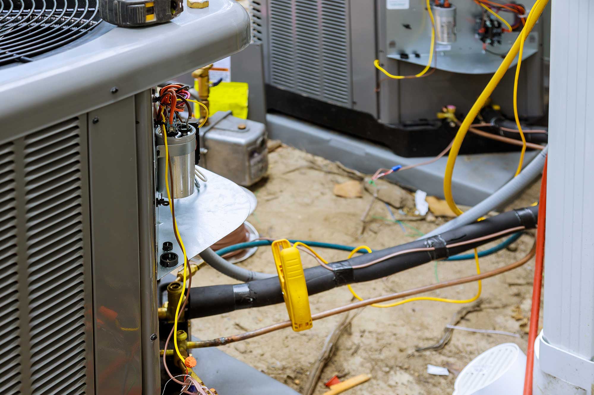 Close-up of an air conditioning unit with exposed wiring and yellow wires. Nearby units are visible, with a sandy ground surface below.