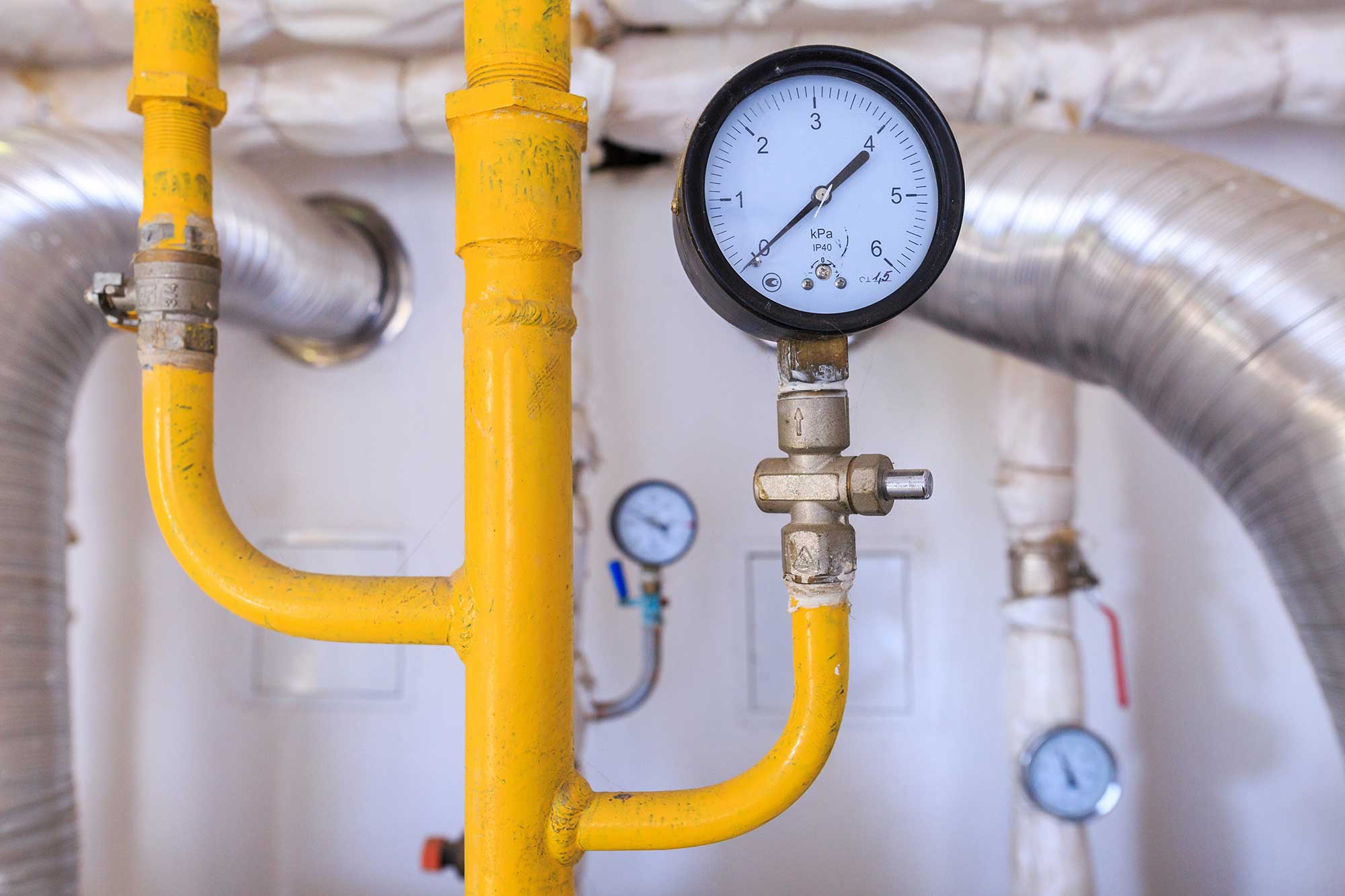 Close-up of a pressure gauge on a yellow pipe in an industrial setting. Metallic ducts surround the main gauge, suggesting a technical environment.