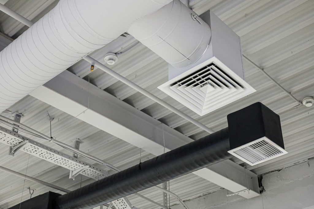 Ceiling air ducts and vents in an industrial or commercial building with exposed pipes and metal beams.